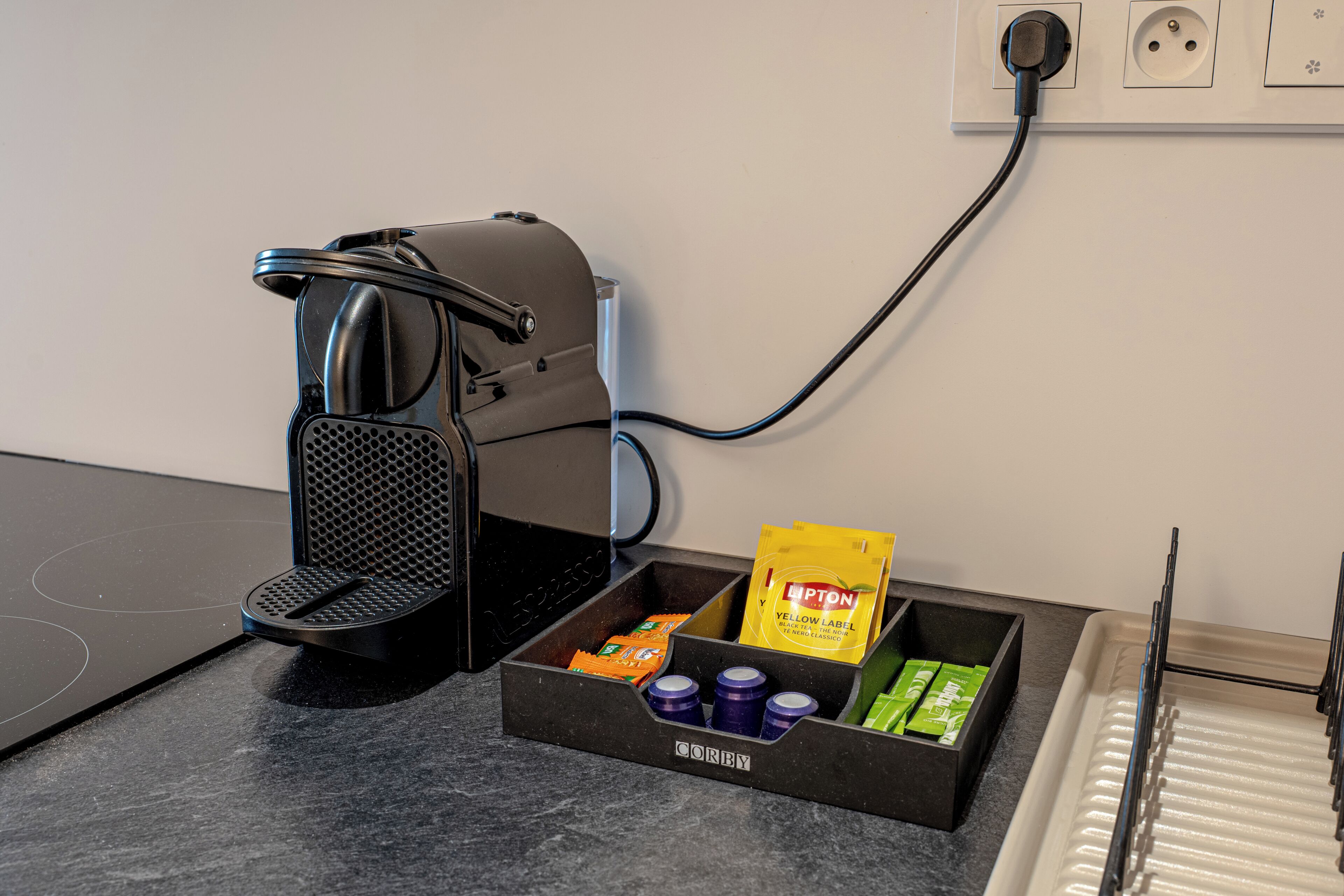 A coffee maker connected to a power outlet, a drawer with coffee pods and a yellow packet, and a metal rack with a tray.