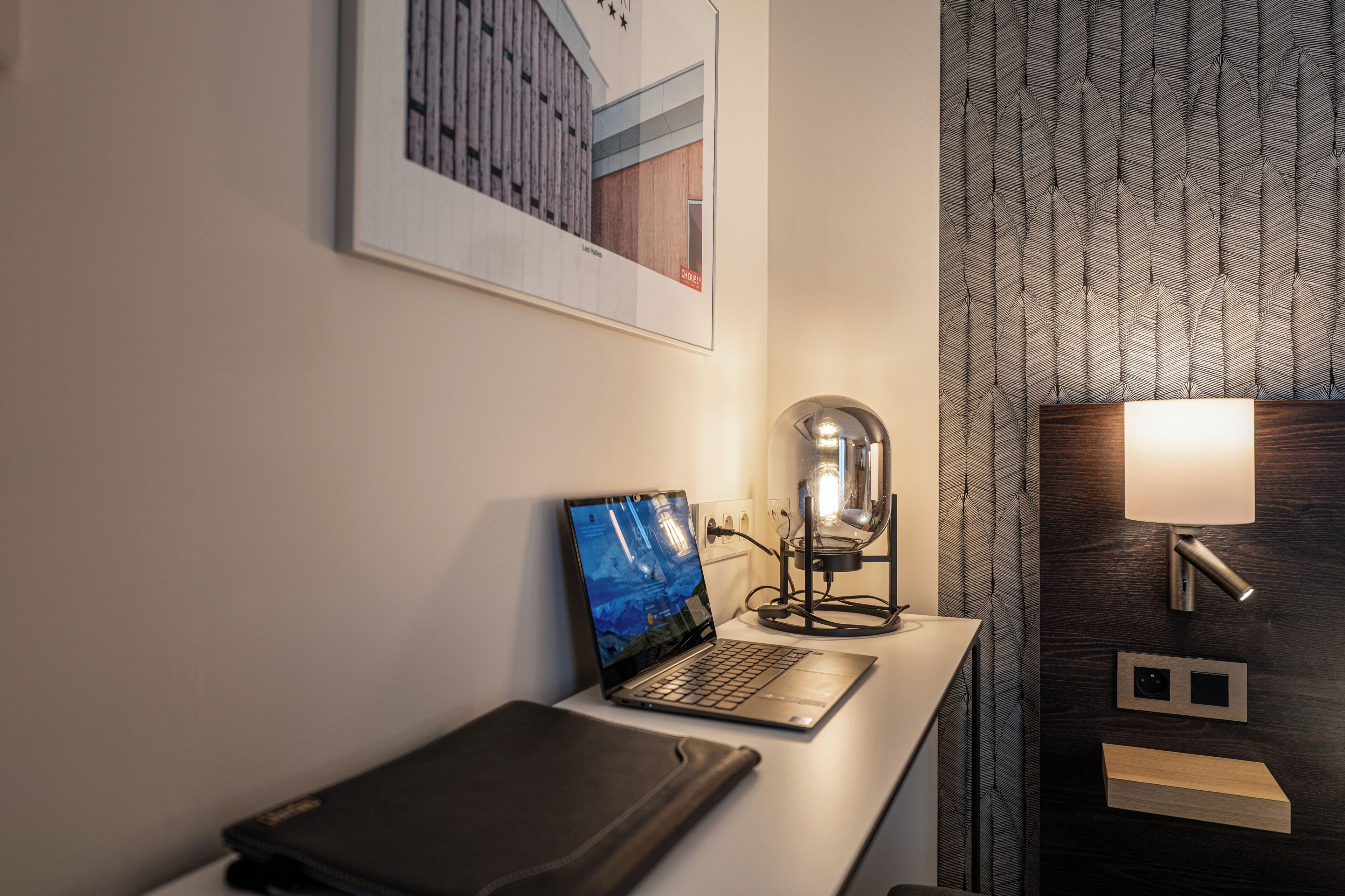 Et moderne hotelværelse med en bærbar computer på et skrivebord, en lampe og et billede i ramme på væggen.