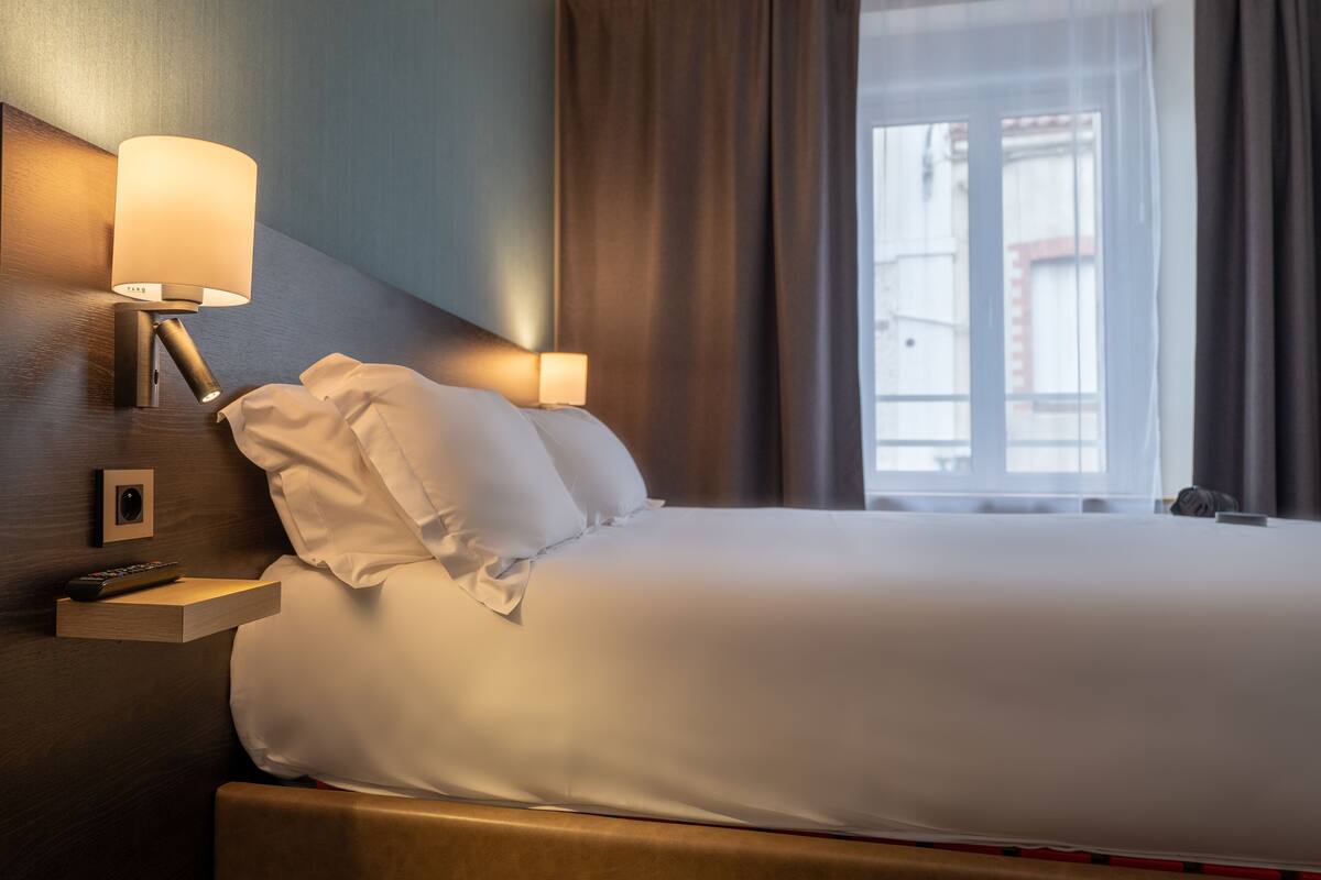 Un lit bien fait, avec du linge de lit blanc, une table de chevet avec une lampe et une télécommande.
