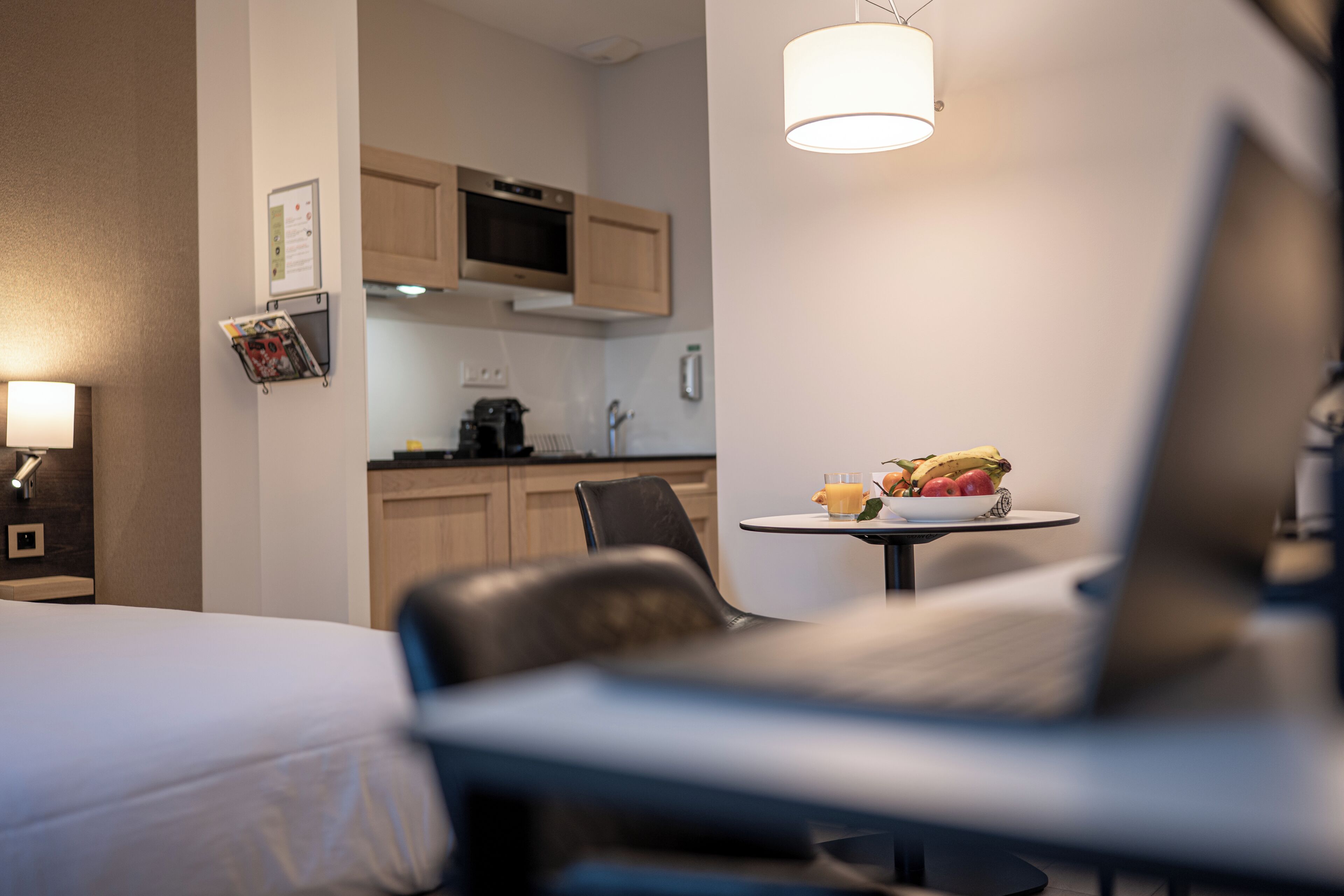 A hotel room with a bed, a desk, a chair, a small table with fruit, a microwave, and a hanging light fixture.