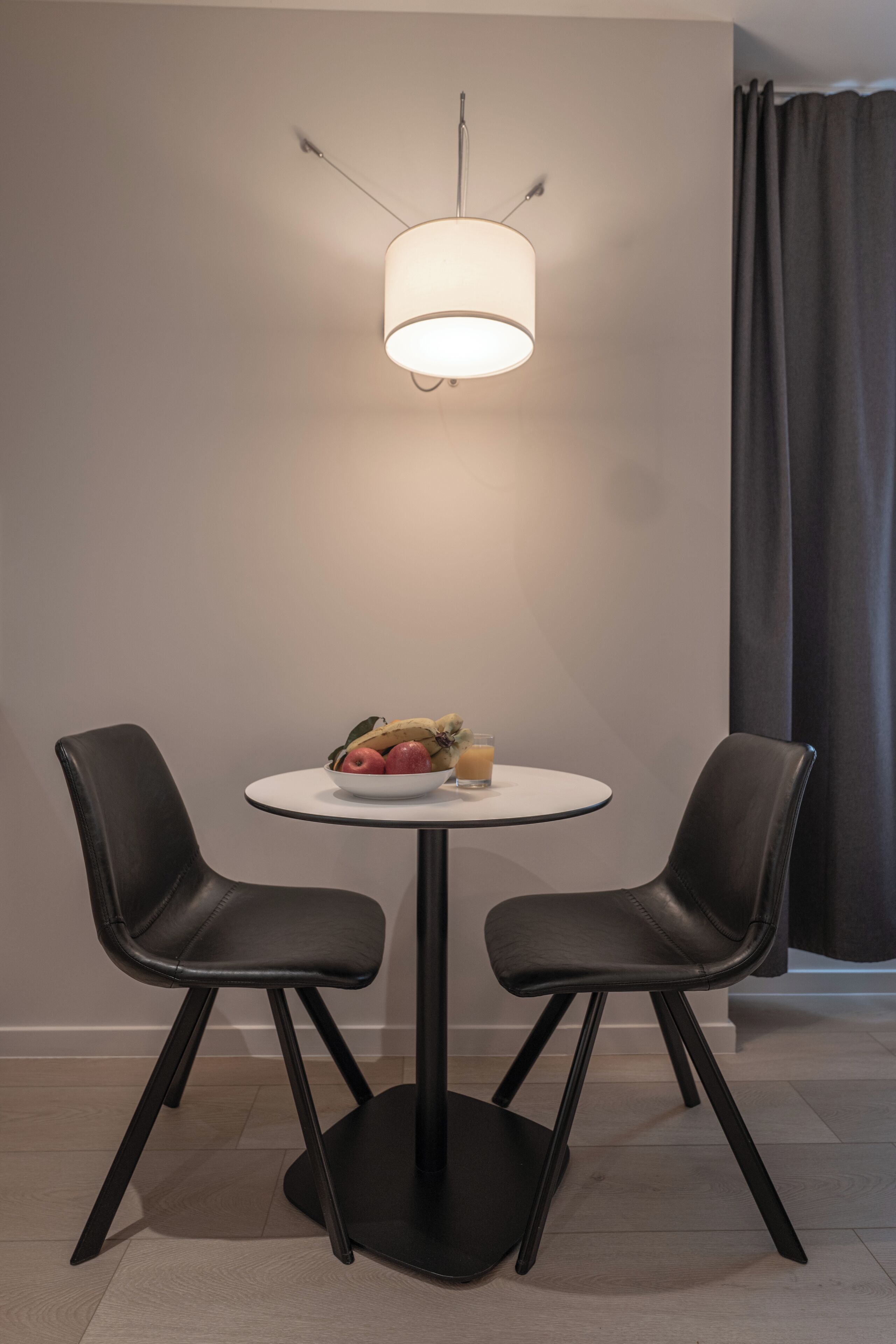 Una habitación moderna con una mesa redonda, dos sillas negras, un bol blanco con frutas y una lámpara colgante.