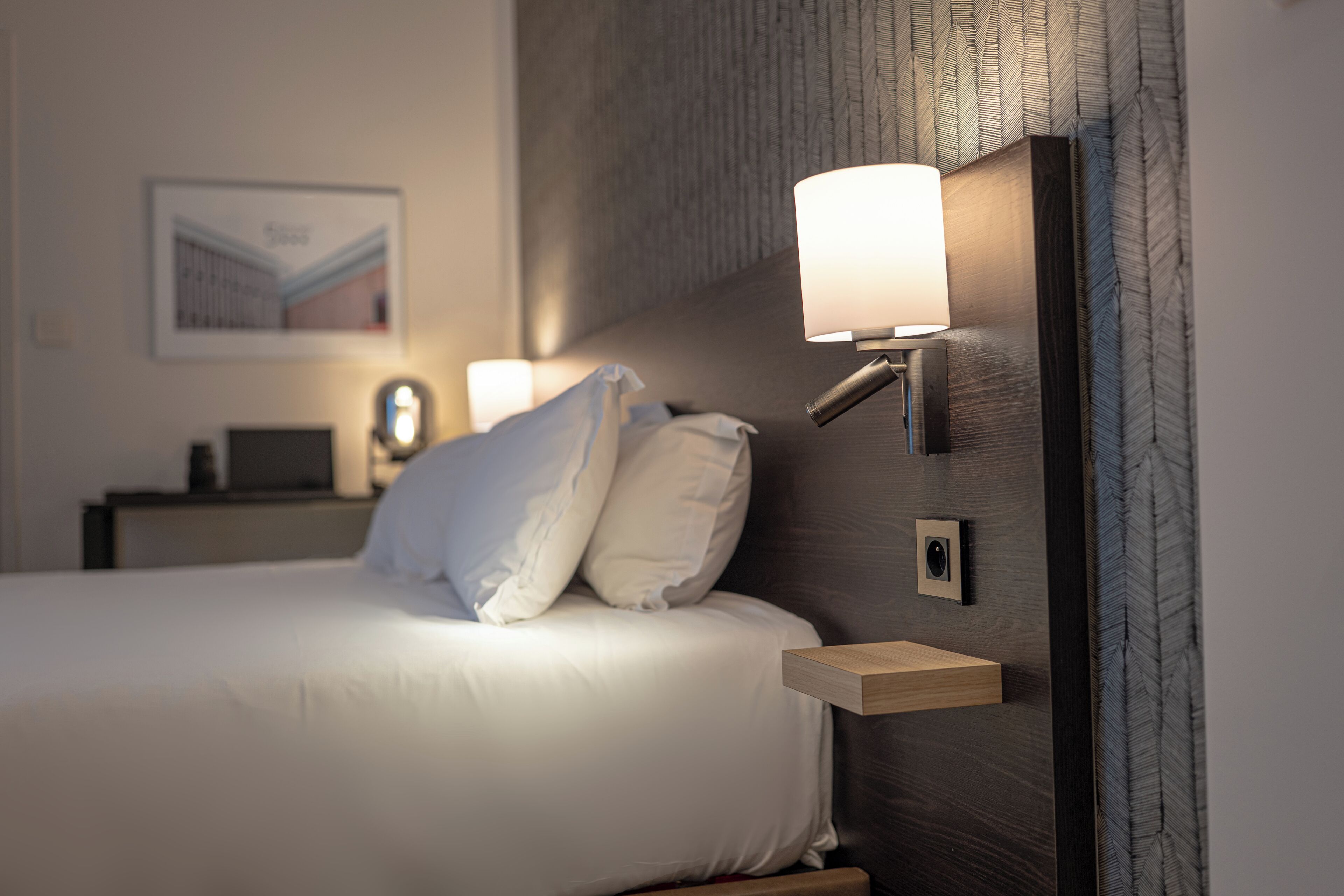 A neatly made bed with white linens, a bedside table with a lamp, and a framed picture on the wall.