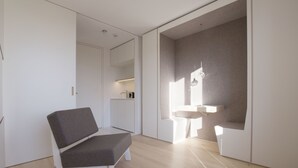 Een moderne, minimalistisch ingerichte kamer met een grijs gestoffeerde bank, een wit wastafelmeubel en een groot raam voor natuurlijk licht.