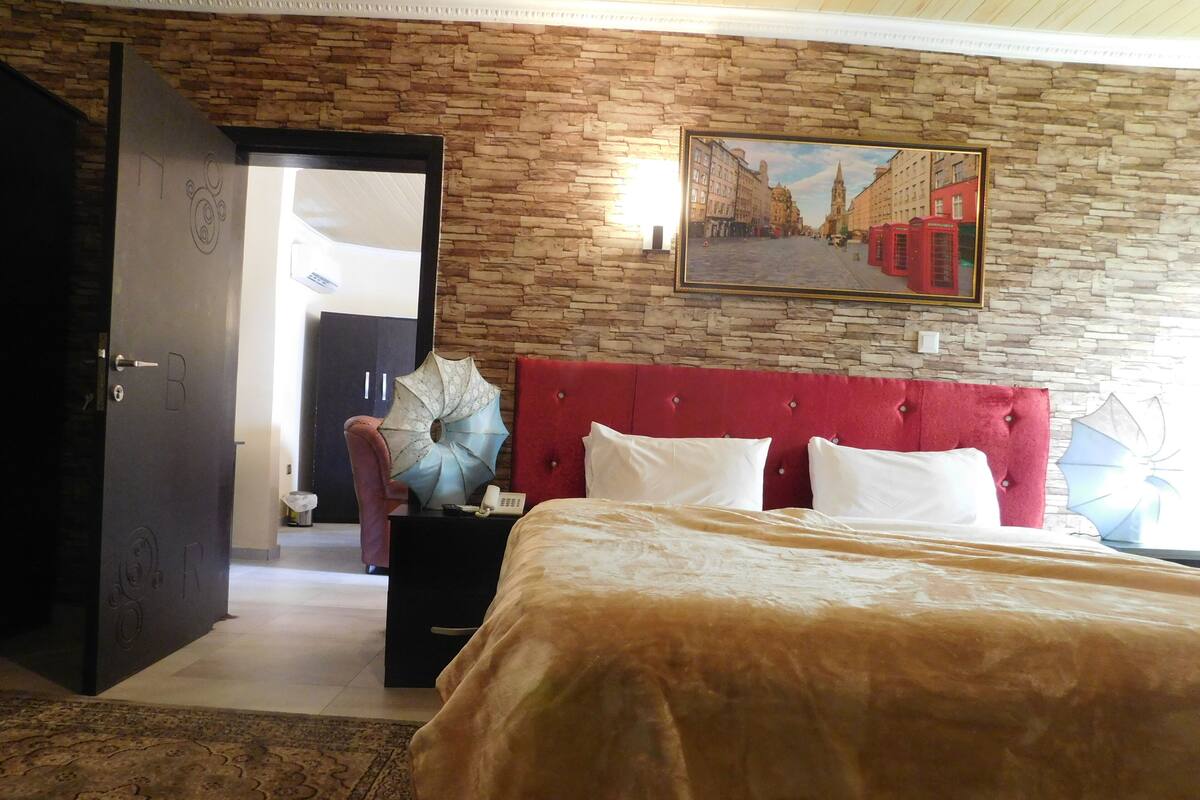 Hotelkamer met een bed, een rood hoofdeinde, een schilderij van een stadsgezicht en een deur met een nummer.