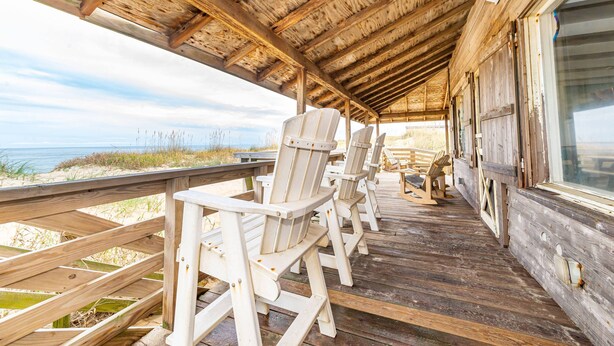 Een houten terras met Adirondack-stoelen en een houten leuning met uitzicht op een strand.