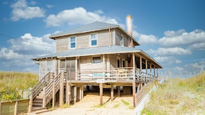Een houten huis met veranda en trap, gelegen op een zandstrand met gras en duinen op de achtergrond.