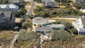 Vista aérea de uma área residencial costeira com várias casas, incluindo uma casa amarela destacada, com varanda e escadaria que leva ao segundo andar.