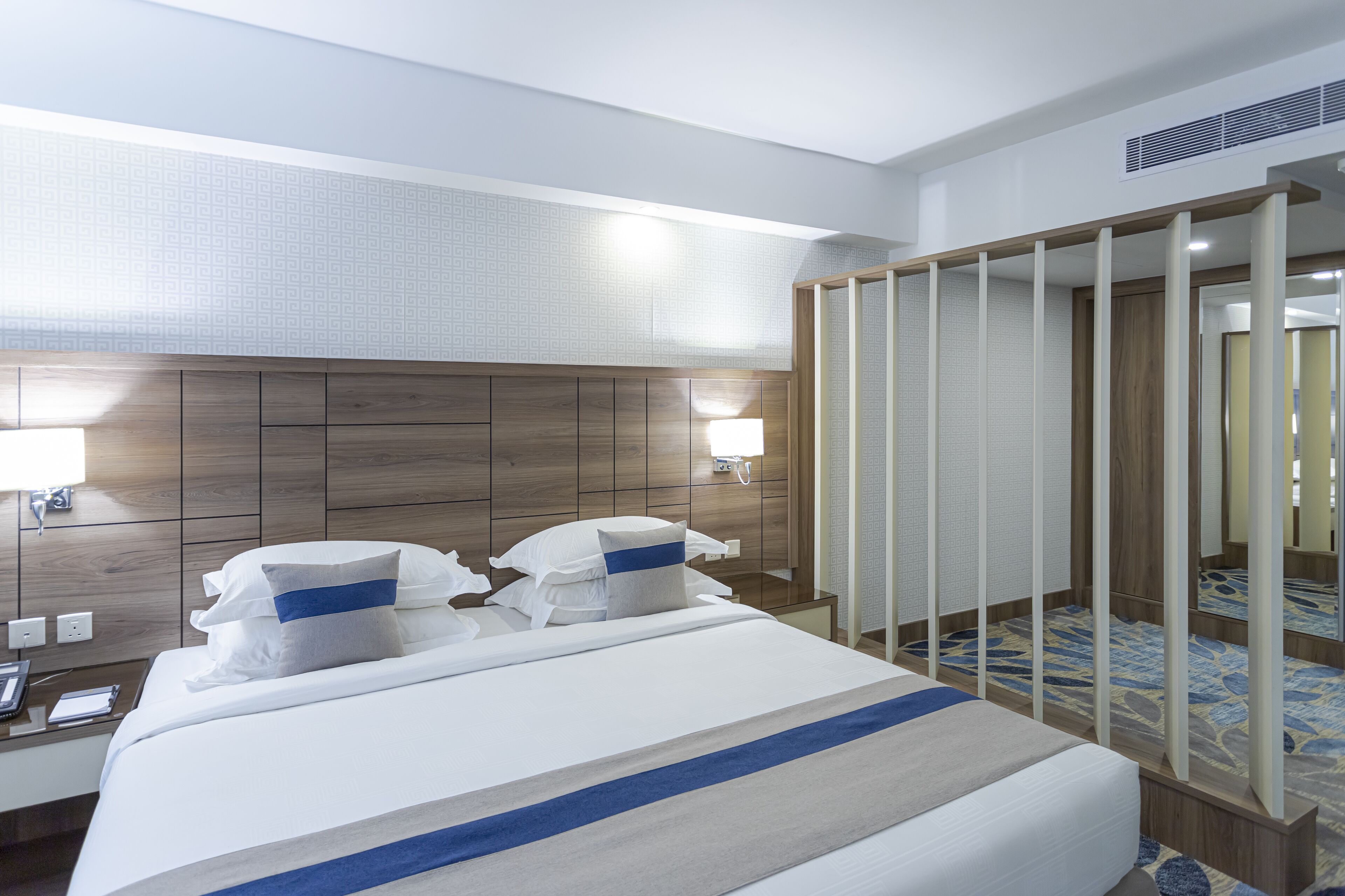 Habitación de hotel con una cama grande, cabecera de madera, mesita de noche, espejo y alfombra estampada.
