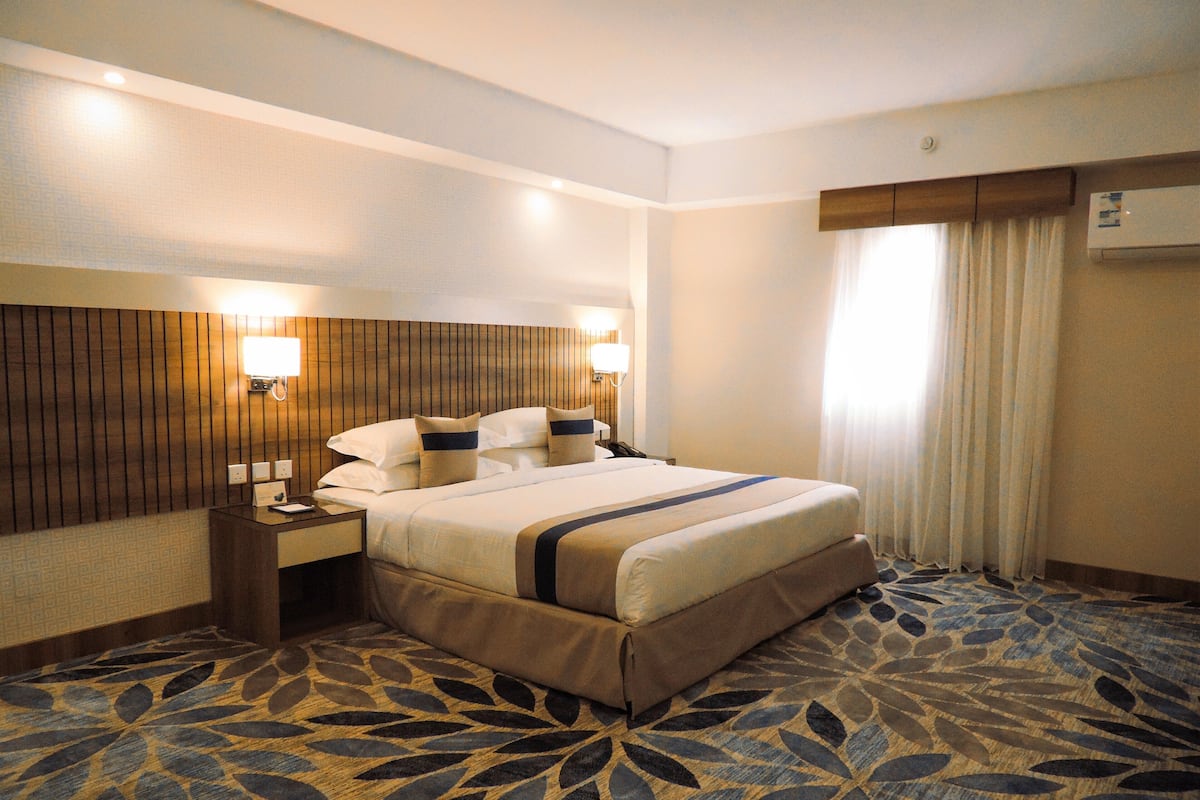 A hotel room with a large bed, wooden headboard, two bedside tables, a nightstand, a window with curtains, and a patterned carpet.