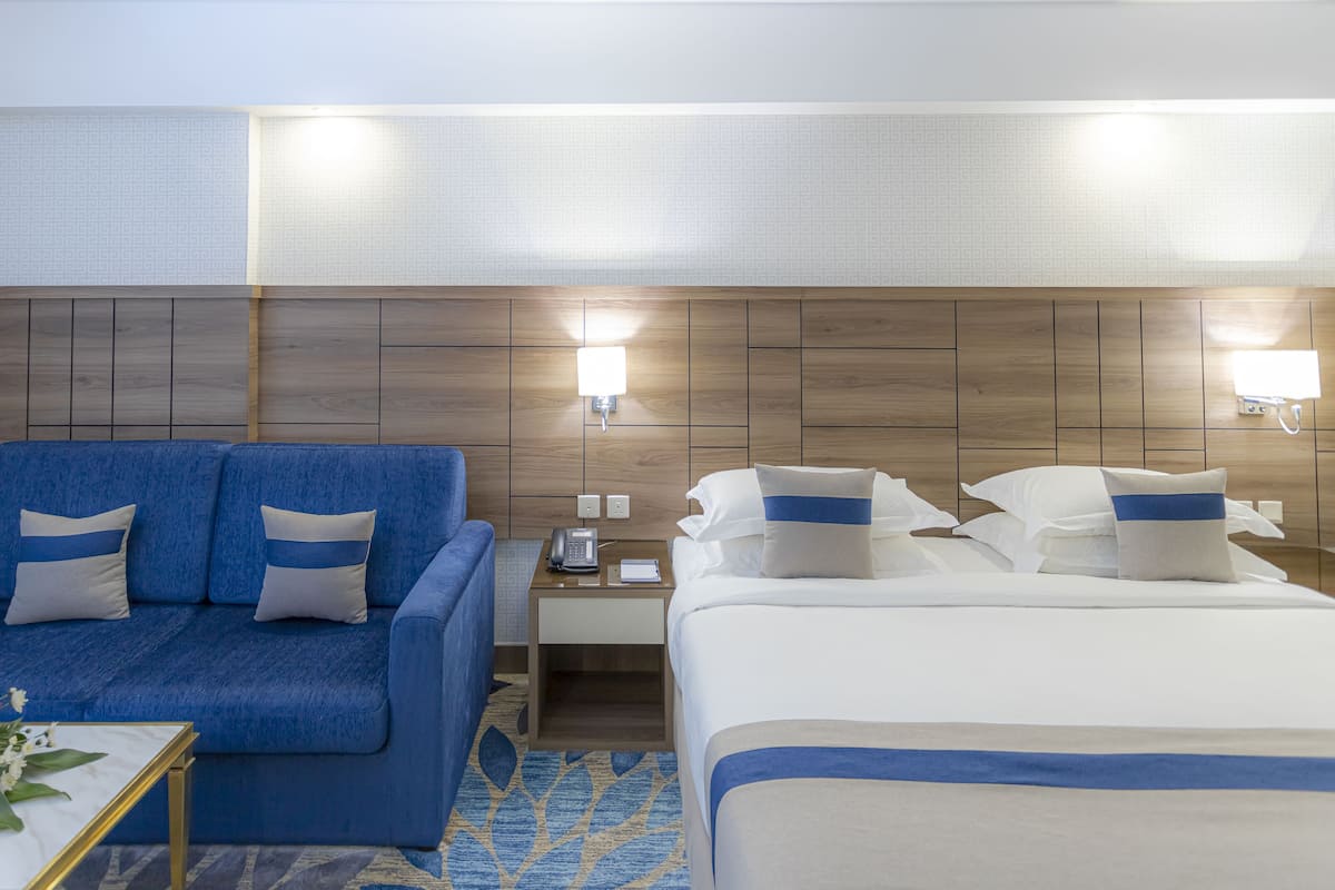 A hotel room with a blue sofa, a bed, a nightstand with a phone, and a lamp.