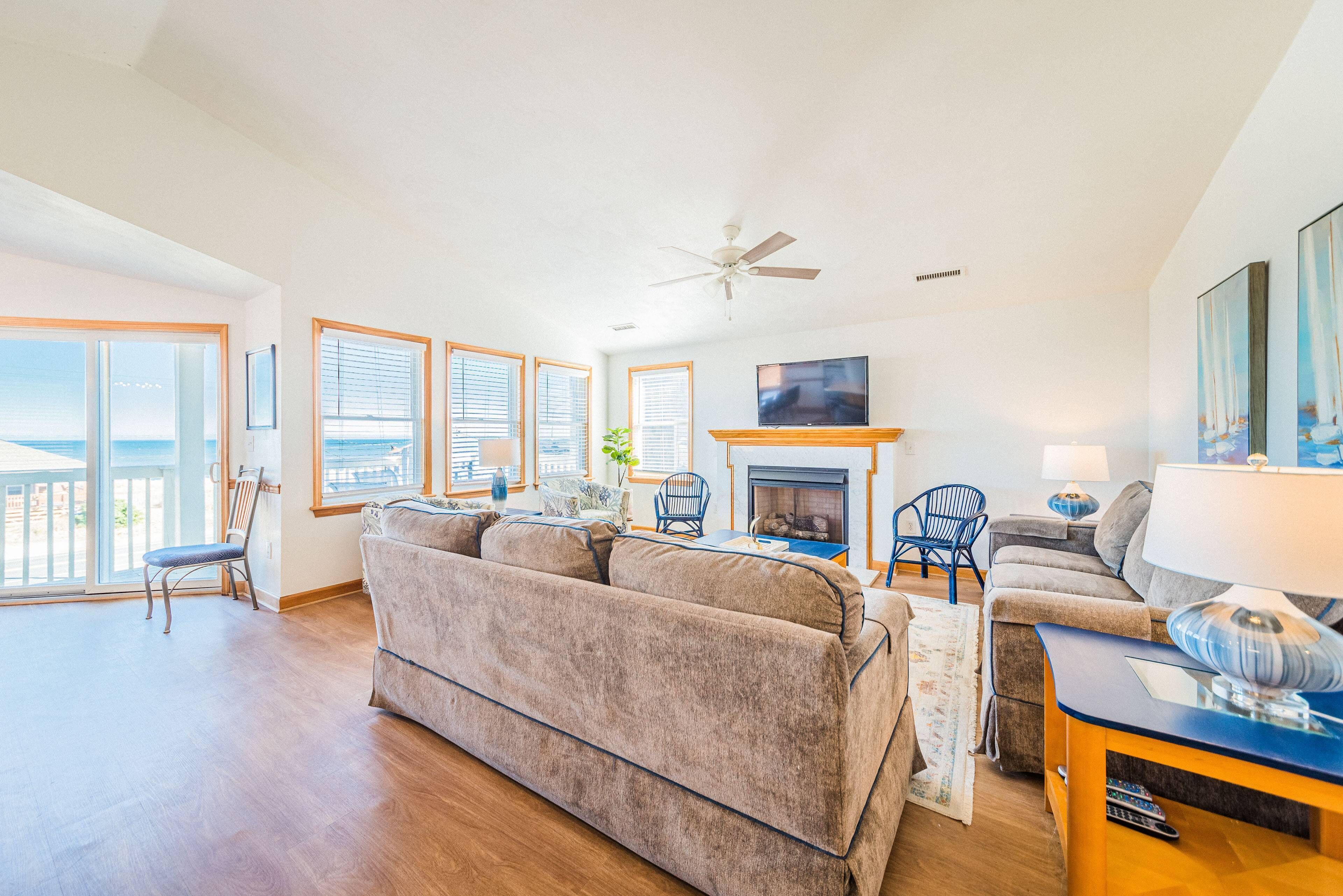 A spacious living room with a large sofa, a television, and a fireplace. There are multiple windows providing natural light and a view of the outdoors.
