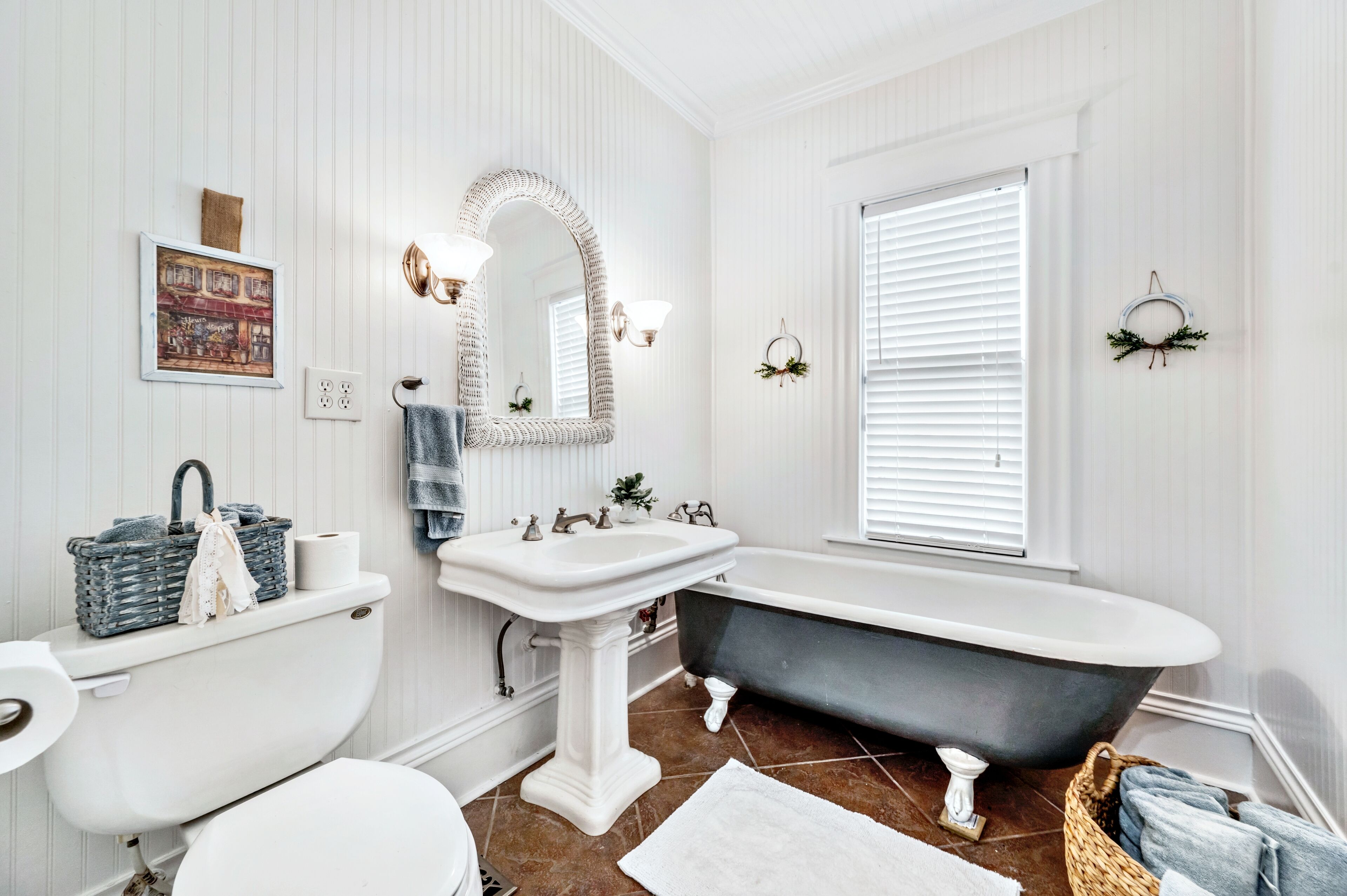 A bathroom with a white toilet, a white sink, and a black freestanding bathtub. There is a mirror above the sink, a window with blinds, and a framed picture on the wall.