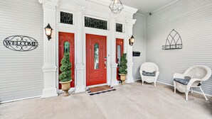 Un porche avec une porte rouge, des colonnes blanches et deux fauteuils en osier.