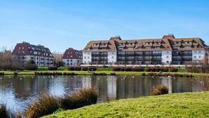 A modern building with multiple balconies, a pond with swans, and a clear blue sky.
