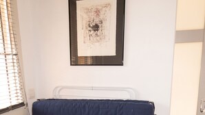 A room with a bed, a window with blinds, a framed artwork on the wall, and a door.