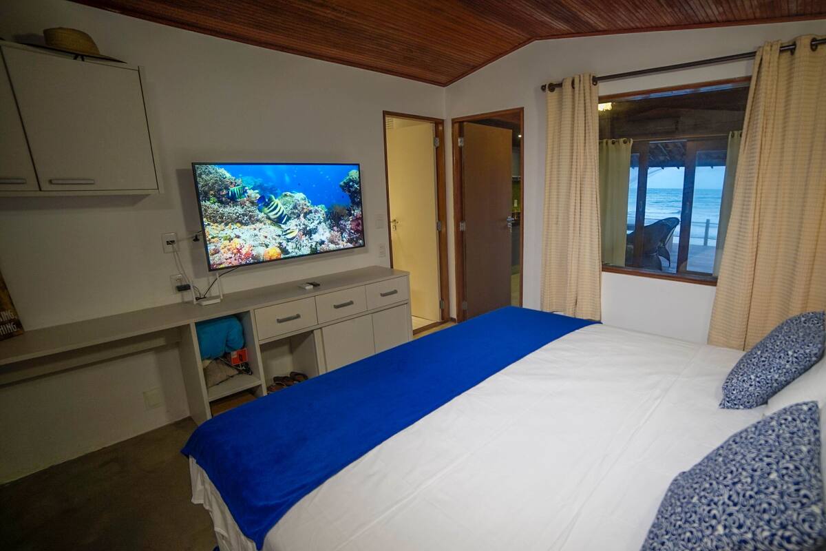 A bedroom with a bed, a TV on a dresser, a window with curtains, and a view of the ocean.