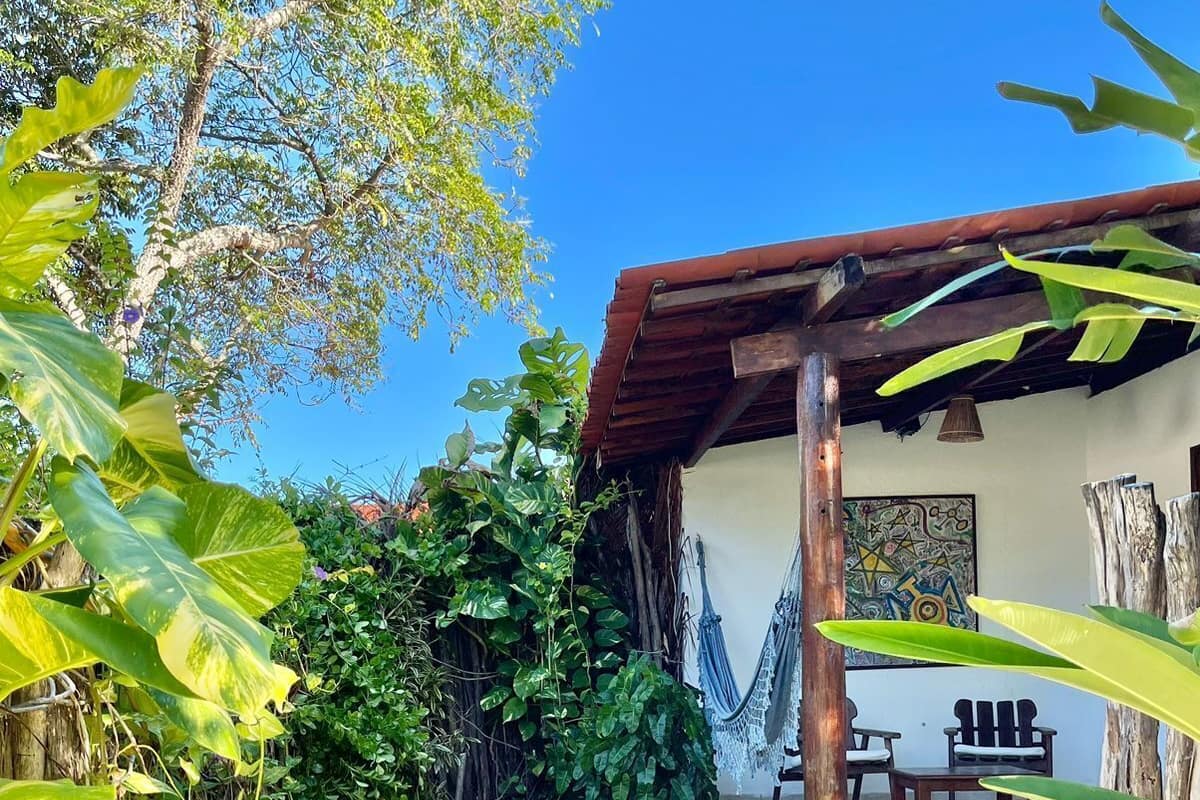 A covered outdoor seating area with a table and chairs, surrounded by lush greenery and a clear blue sky.