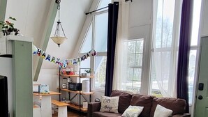 A spacious living room with a high ceiling, large windows, a brown sofa, a wooden coffee table, and a TV on the left.