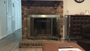 Een woonkamer met een open haard, een televisie erboven, een klok aan de muur, en een leren voetenbankje met een afstandsbediening erop.
