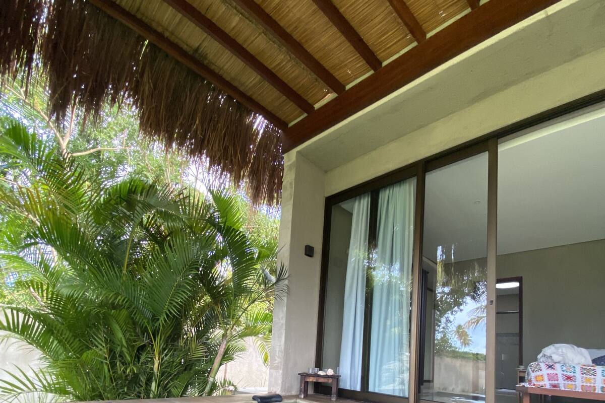 Une piscine dotée d’une terrasse carrelée, entourée d’une végétation luxuriante et surmontée d’une structure à toit de chaume.