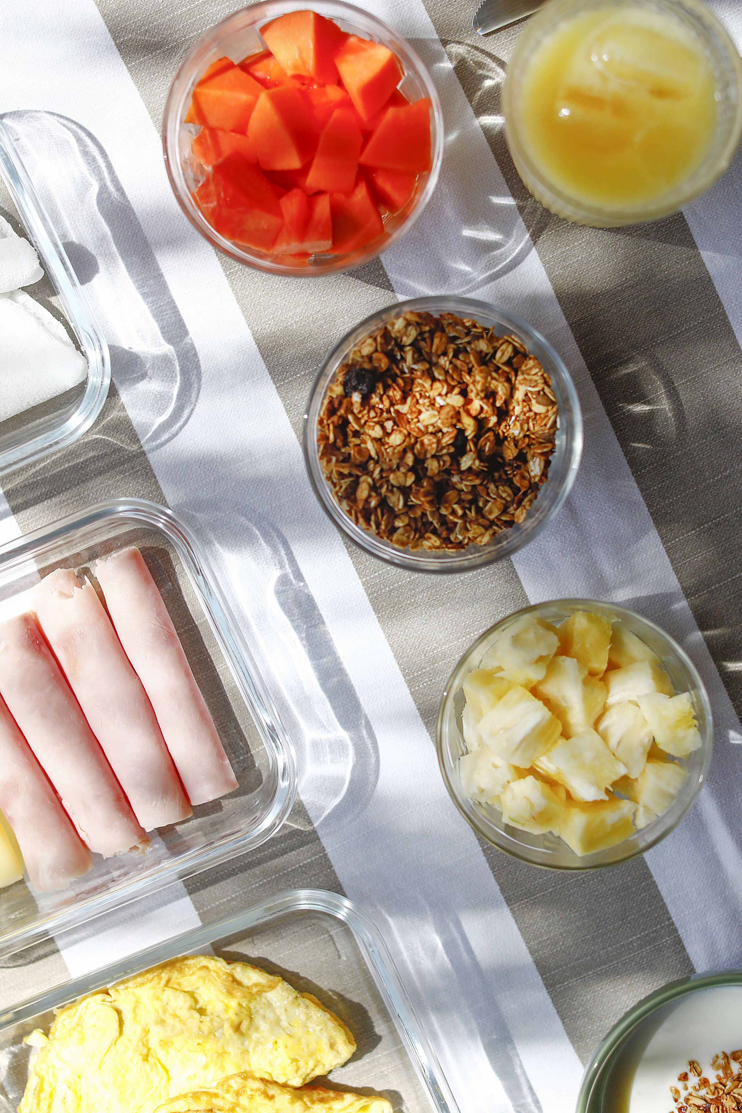 Une table où sont présentés divers aliments, notamment du jambon, de l’ananas, du melon, du granola et un yaourt en parfait.