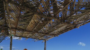 A beach umbrella structure with a wooden frame and thatched roof, situated on a sandy beach with clear blue sky and ocean in the background.