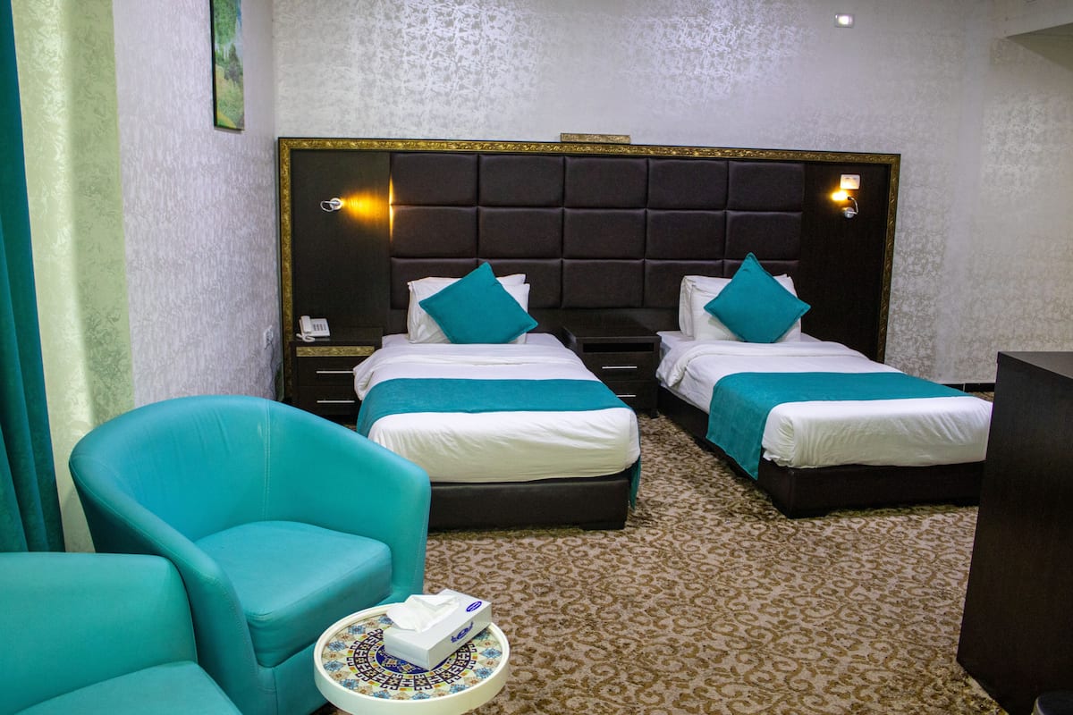 A hotel room with two beds, a teal armchair, a bedside table with a lamp, and a patterned carpet.