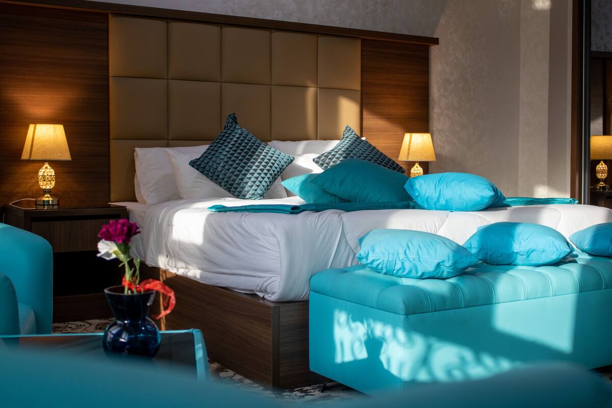 A modern bedroom with a large bed, two bedside tables with lamps, a blue armchair, and a vase with flowers.