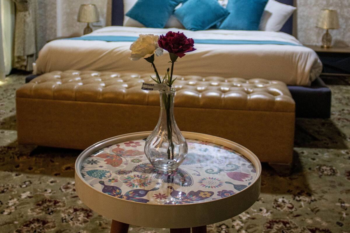 A hotel room with a bed, a nightstand, a lamp, a round table with a vase of flowers, and a patterned carpet.