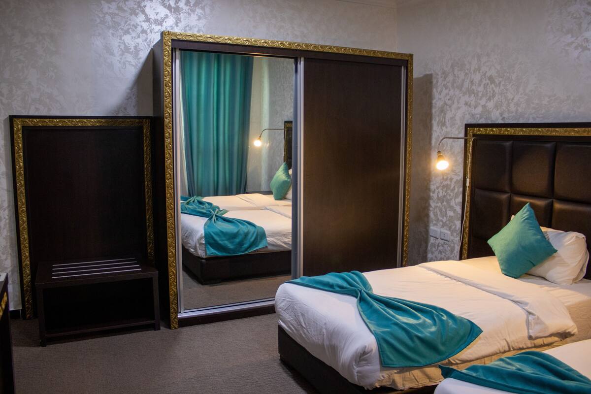 A hotel room with a large mirror, a bed with teal bedding, a nightstand, and a wall-mounted lamp.