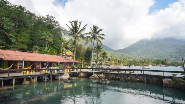 A wooden bridge over calm water, a building with a red roof, and lush greenery in the background. - Aquário Pousada Hostel (Angra dos Reis)