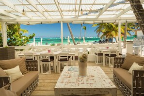 An outdoor seating area with white furniture, a glass table with a decorative item, and a view of the beach and ocean. - Costa Hermosa C302LO_200Mpbs_Pool_BBQ_Walk to the Beach (Bavaro-Punta Cana)