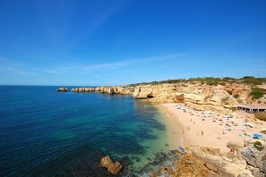 A coastal beach with clear blue waters, rocky cliffs, and a sandy shore with umbrellas and people.