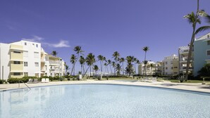 A resort pool area with clear blue water, palm trees, and modern buildings. - Playa Turquesa I201 LO_200 Mbps_Private Beach Access_BBQ (Bavaro-Punta Cana)