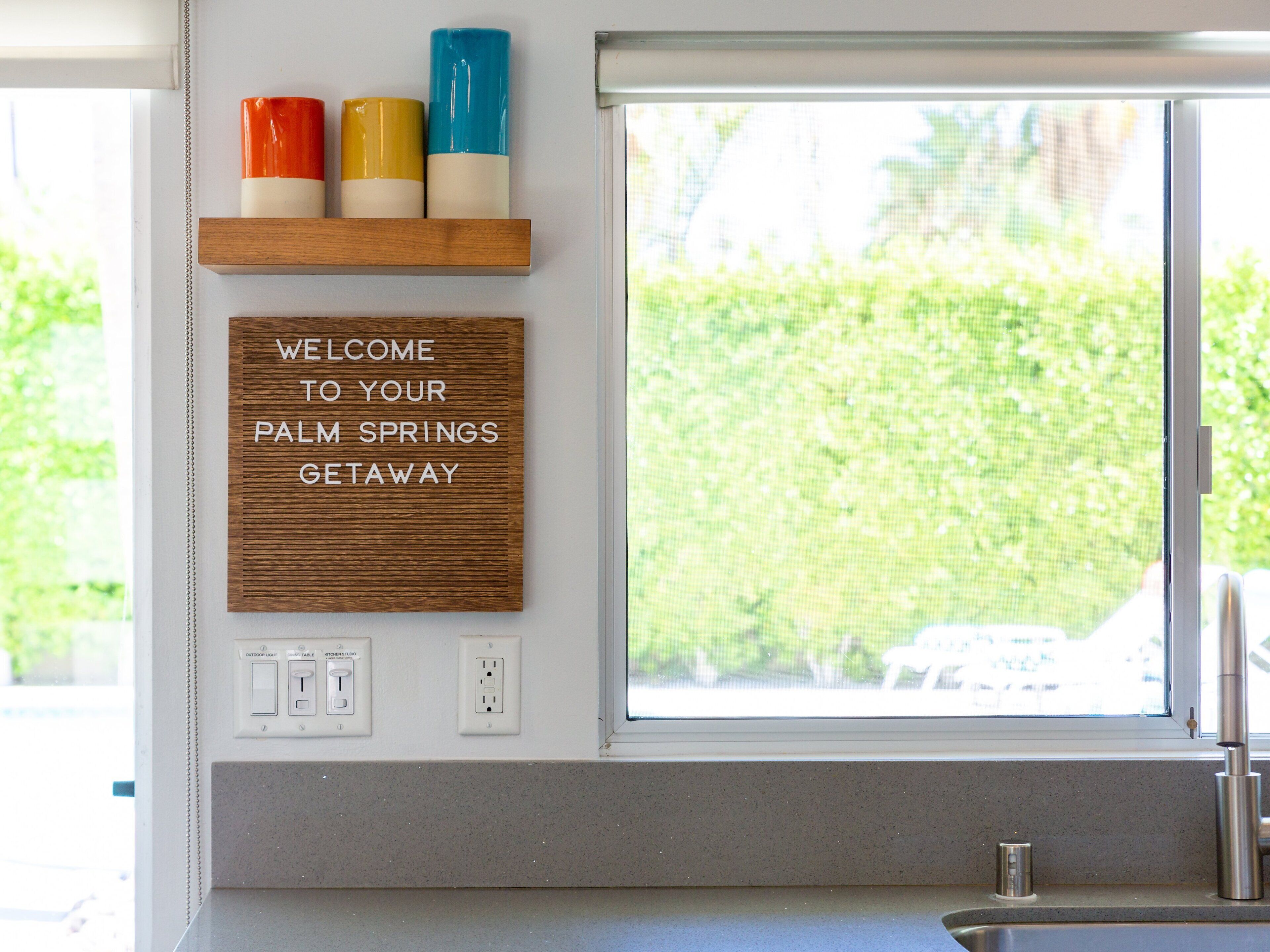 A kitchen with a window, a welcome sign, and a shelf with colorful containers.