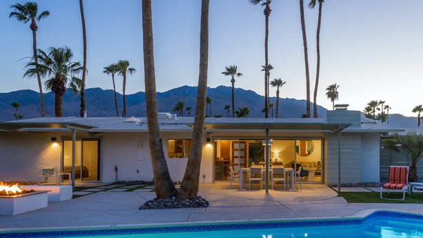 Una casa moderna con piscina, palmeras y montañas al fondo.