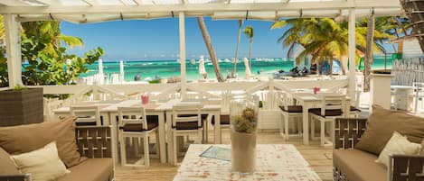 An outdoor seating area with white furniture, a glass table with a decorative item, and a view of the beach and ocean.