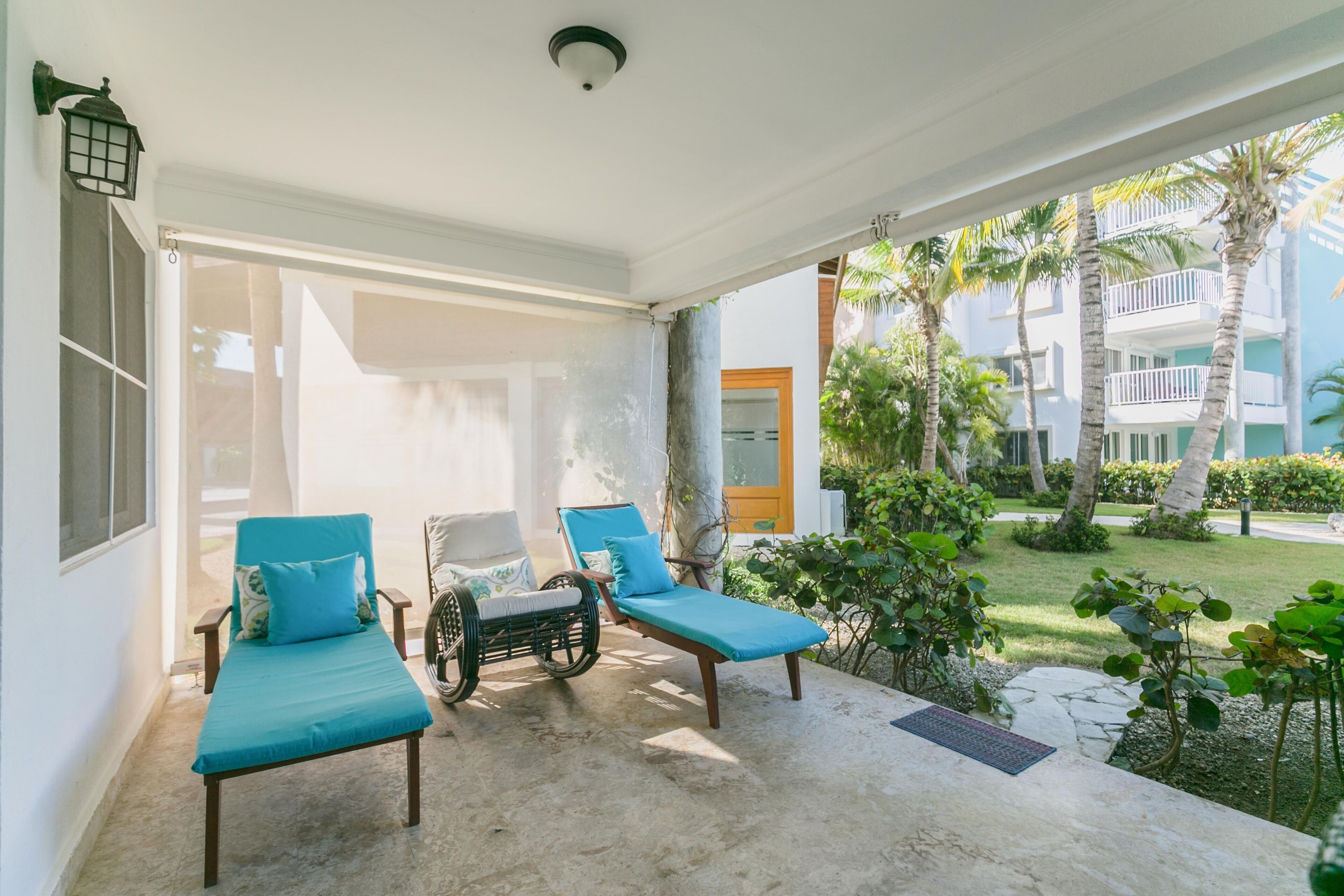 Un espace extérieur aménagé, abrité, avec deux fauteuils de relaxation et un fauteuil en osier, un sol en pierre et une vue sur un immeuble doté de balcons et de palmiers.