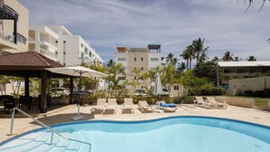 Une piscine entourée de palmiers et d’un bâtiment doté de nombreux balcons, avec des chaises longues et des parasols à proximité.