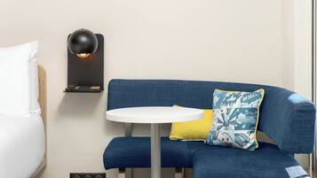 A modern hotel room with a blue sofa, a small round table, a lamp, and a bed with white bedding.