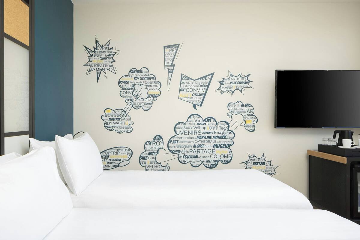 Ein Hotelzimmer mit Bett, einem an der Wand montierten Fernseher und einer Wand mit verschiedenen Wörtern und Sprüchen.