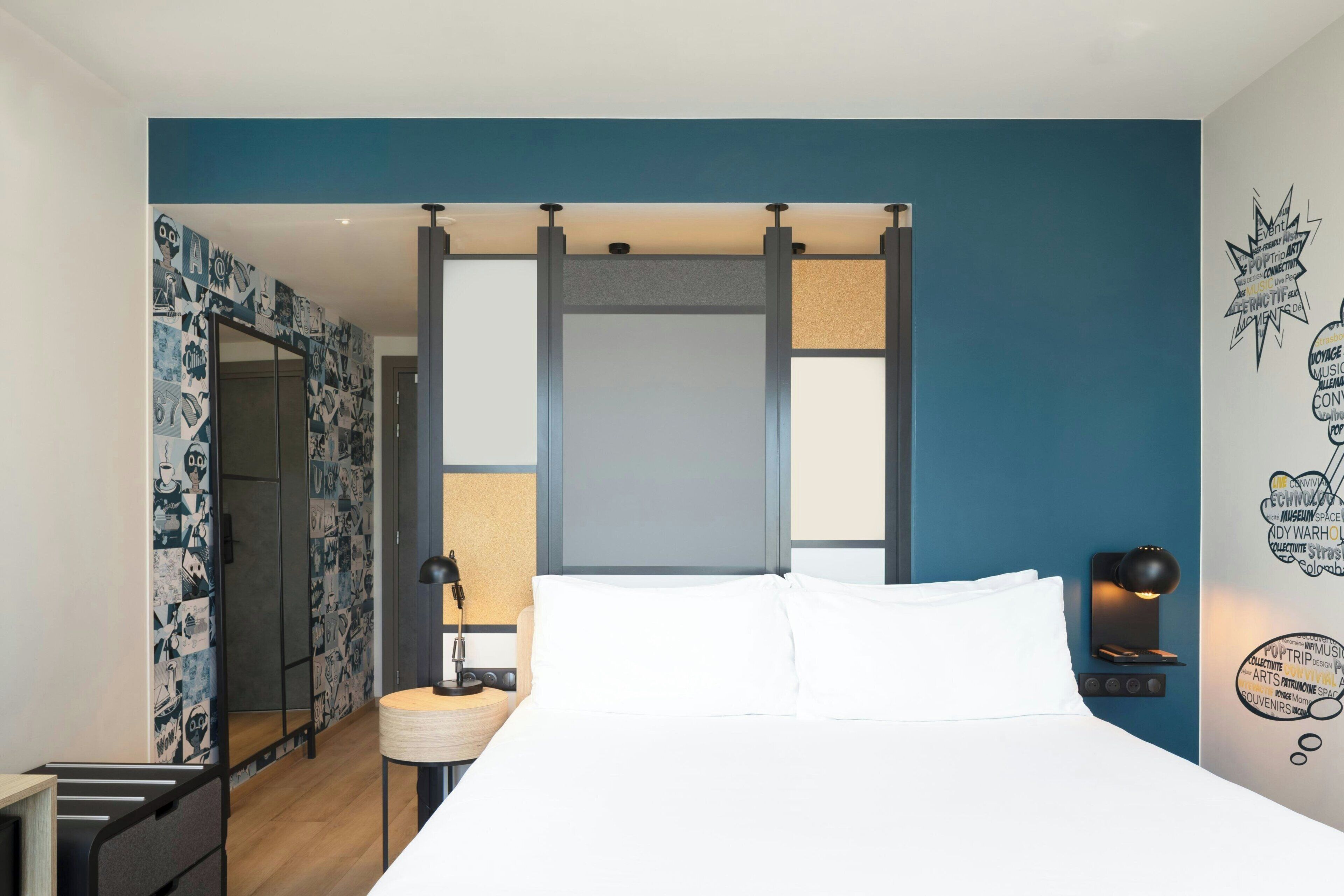 Habitación de hotel con cama, mesita de noche, lámpara, pared con una nube de palabras y puerta que da a un baño.
