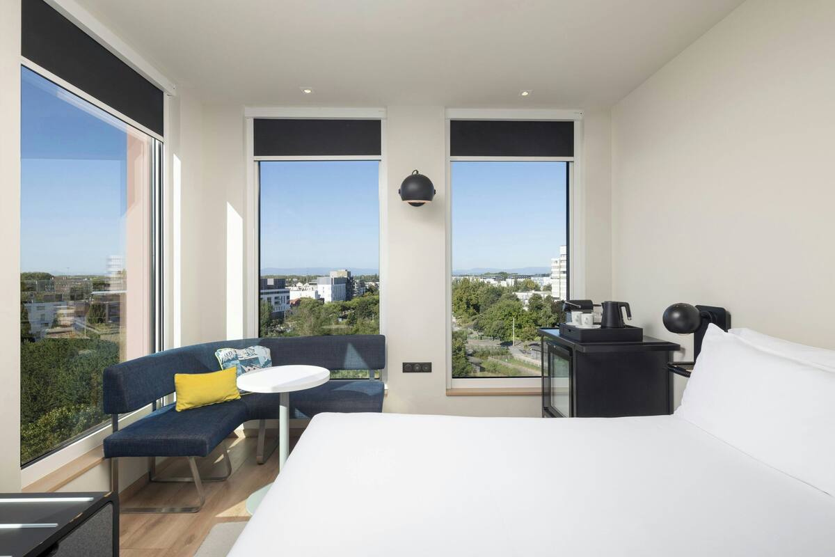 Ein modernes Hotelzimmer mit einem großen Bett, einem kleinen Tisch, einer Couch und einem Schreibtisch mit Lampe. Das Zimmer verfügt über große Fenster mit Blick auf die Stadt.