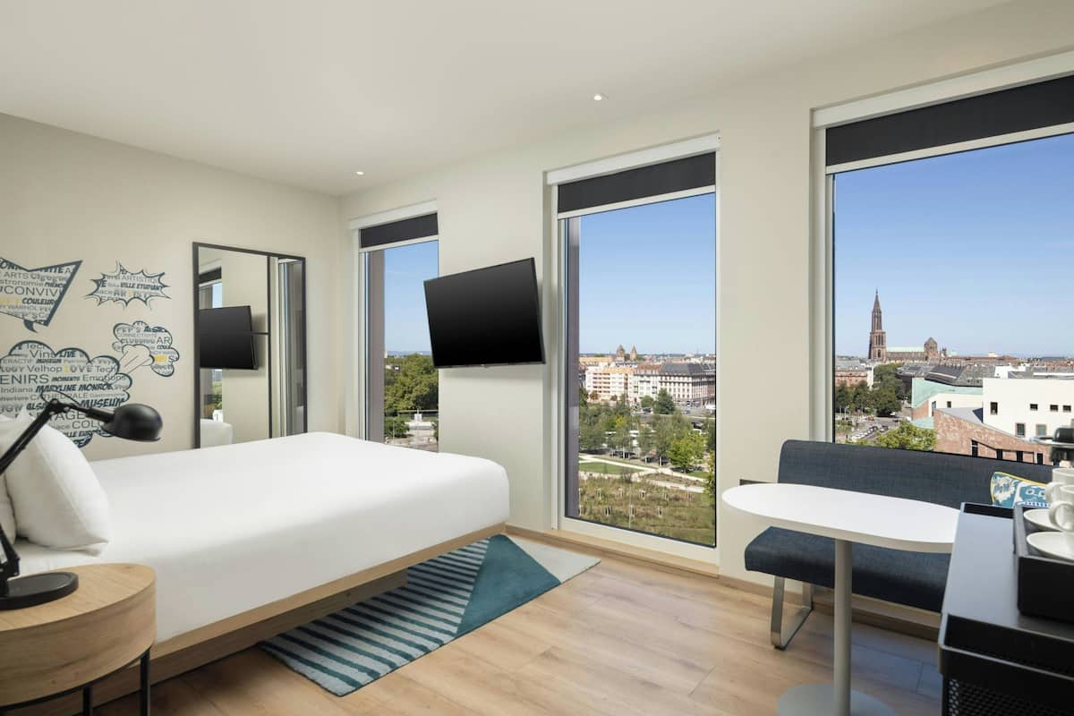 Ein modernes Hotelzimmer mit Bett, Schreibtisch, Stuhl, Fernseher und großen Fenstern mit Blick auf die Stadt.