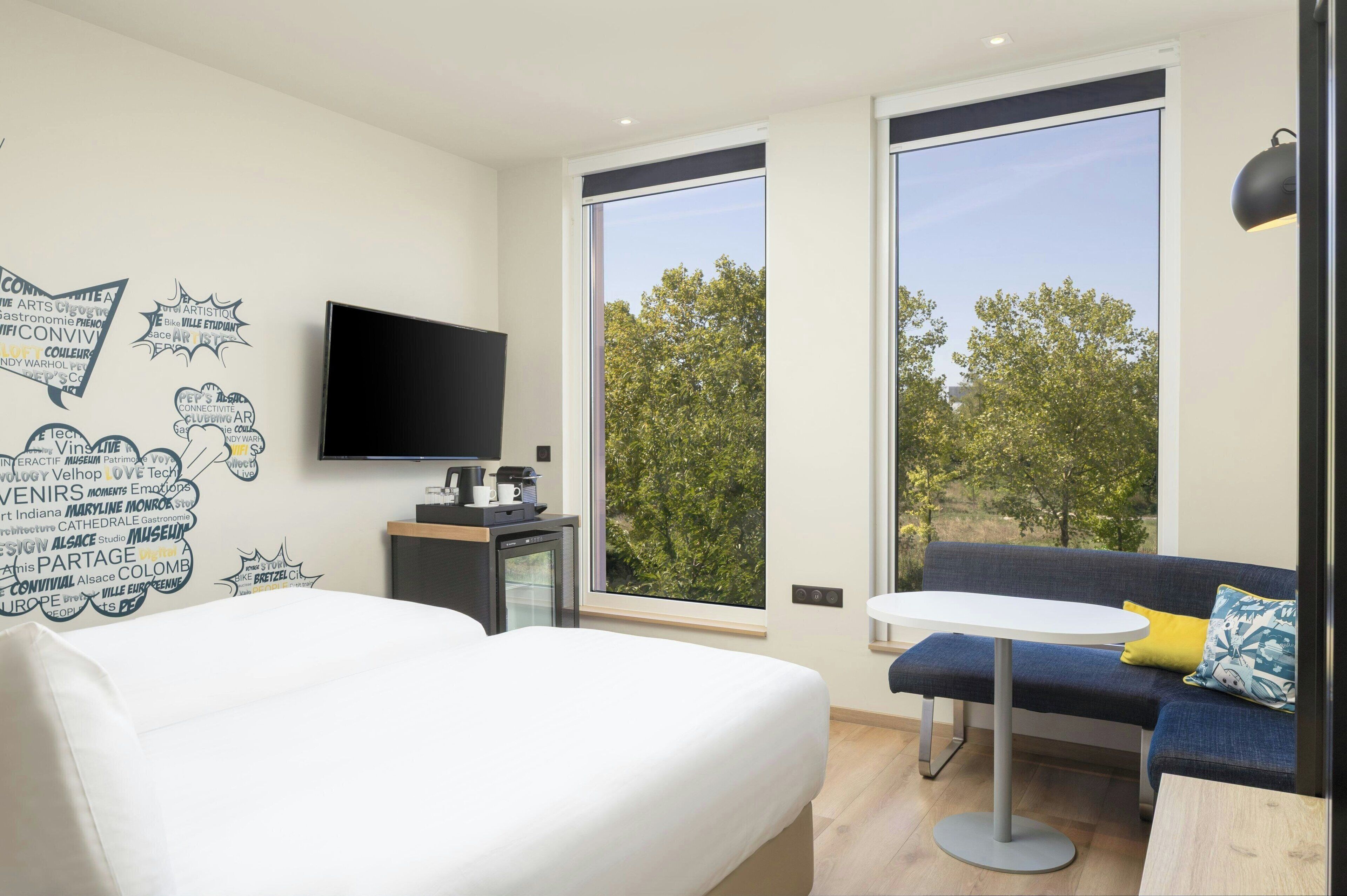 Habitación de hotel moderna con cama, escritorio con televisor de pantalla plana, pequeña mesa y sofá con vistas a árboles a través de amplias ventanas.