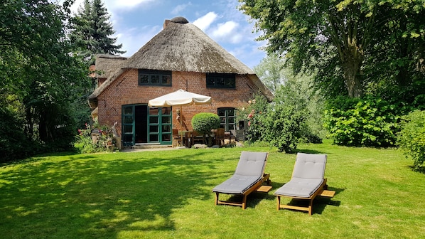 Una casa con techo de paja y fachada de ladrillo, un césped con dos sillas de descanso y una zona de comedor con mobiliario exterior.