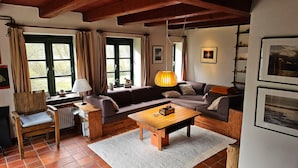 A cozy living room with a sofa, a wooden coffee table, and a chair. There are windows with curtains, a lamp, and framed pictures on the wall.