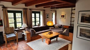 Una sala de estar acogedora con un sofá, una mesa de centro de madera y un sillón. Hay ventanas con cortinas, una lámpara y cuadros enmarcados en la pared.