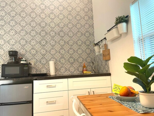 Una cocina con papel tapiz estampado, un microondas, una cafetera, un lavamanos y una mesa de madera con una planta en maceta.