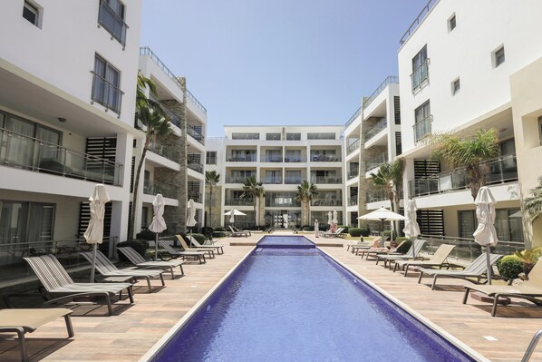 A pool area with lounge chairs and umbrellas, surrounded by multi-story buildings with balconies.